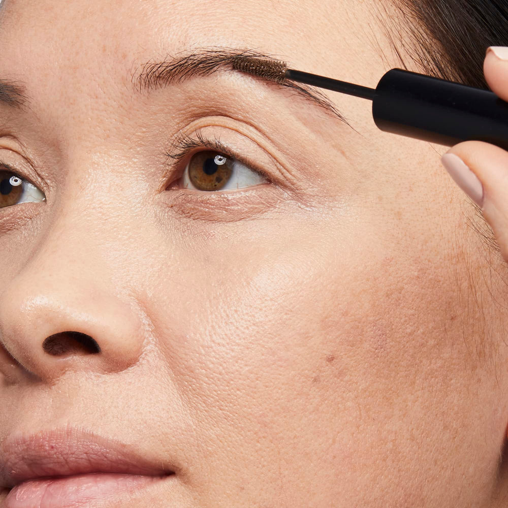 Applying gel to brows with curved wand for fuller shape.
